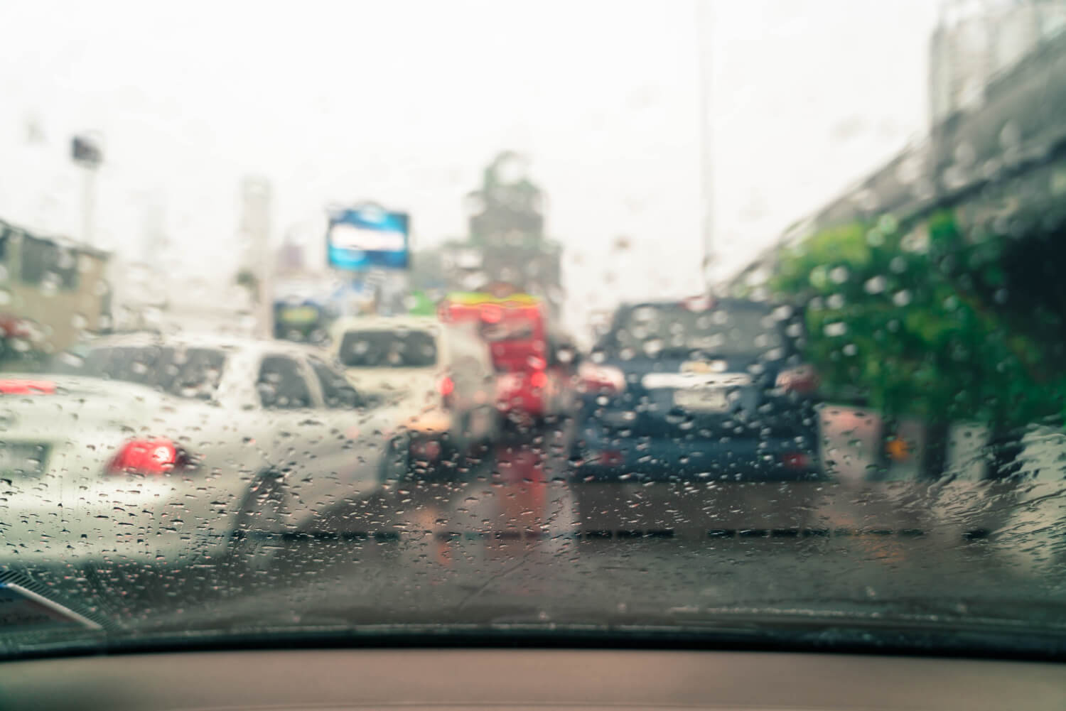 View through a car windshield covered with raindrops, showing blurred traffic on a wet city road during rainy weather.